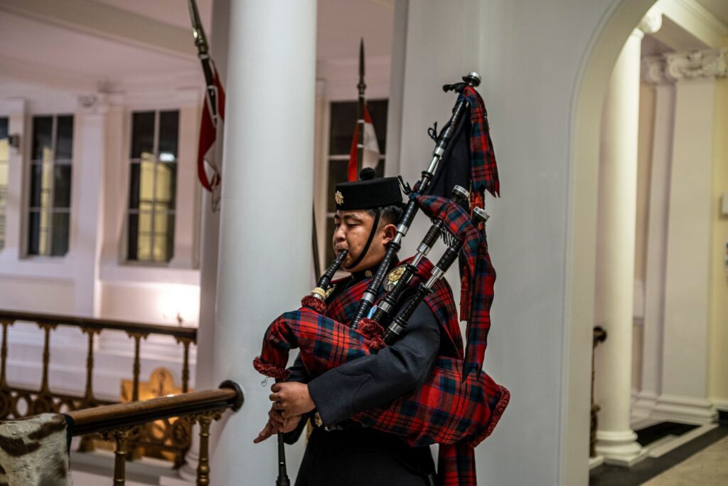 A man wearing traditional Scottish attire plays bagpipes inside a hall.
