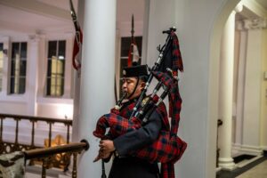 A man wearing traditional Scottish attire plays bagpipes inside a hall.